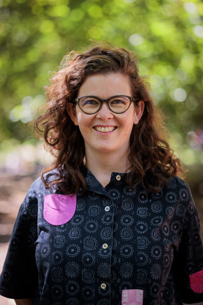 Photo of a white woman smiling, with reddish curly hair and wearing glasses. She seems to be outside in a park, and is wearing a Marimekko top.